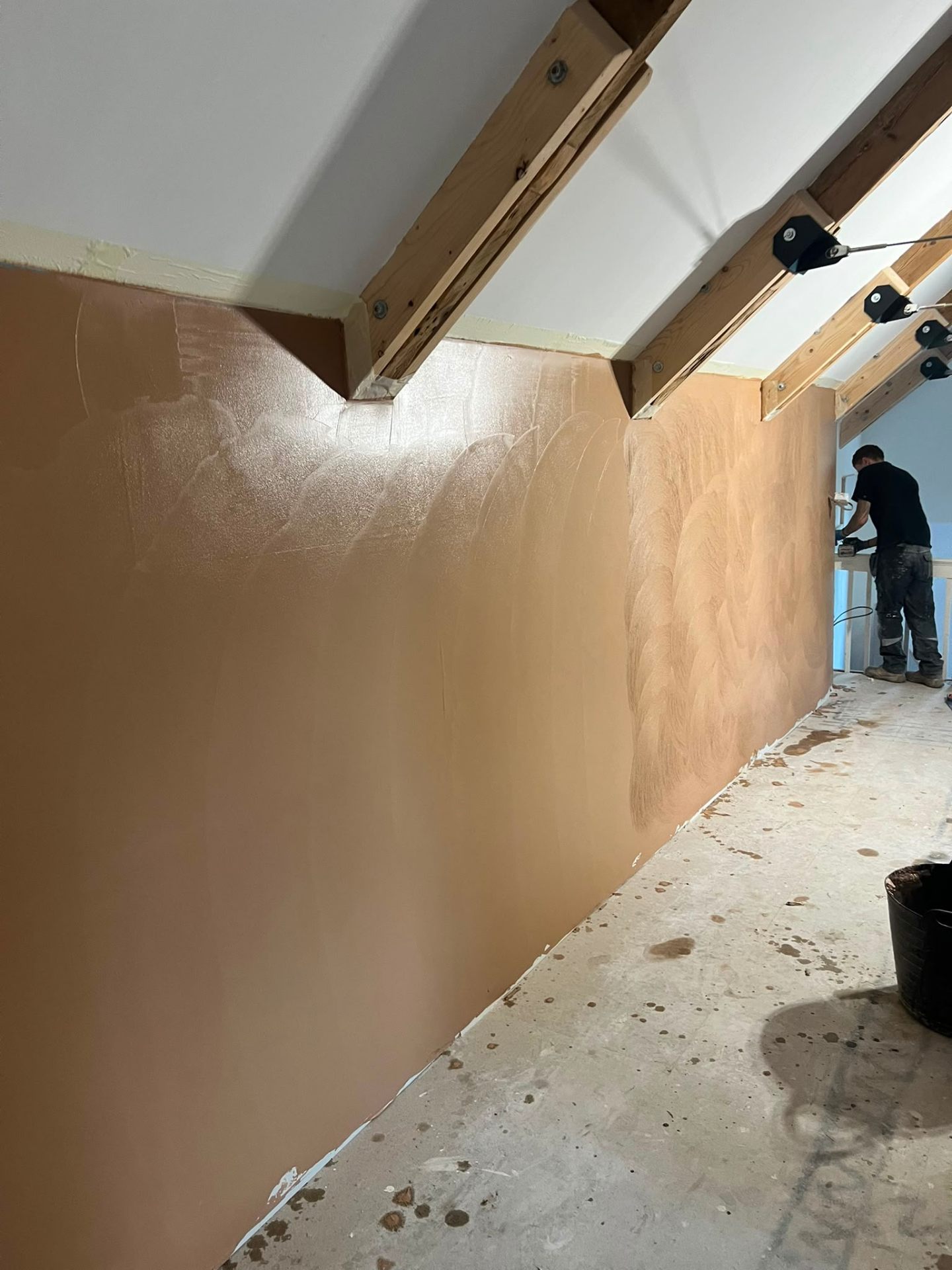 Worker applying fresh brown plaster to an interior wall beneath exposed timber roof rafters.