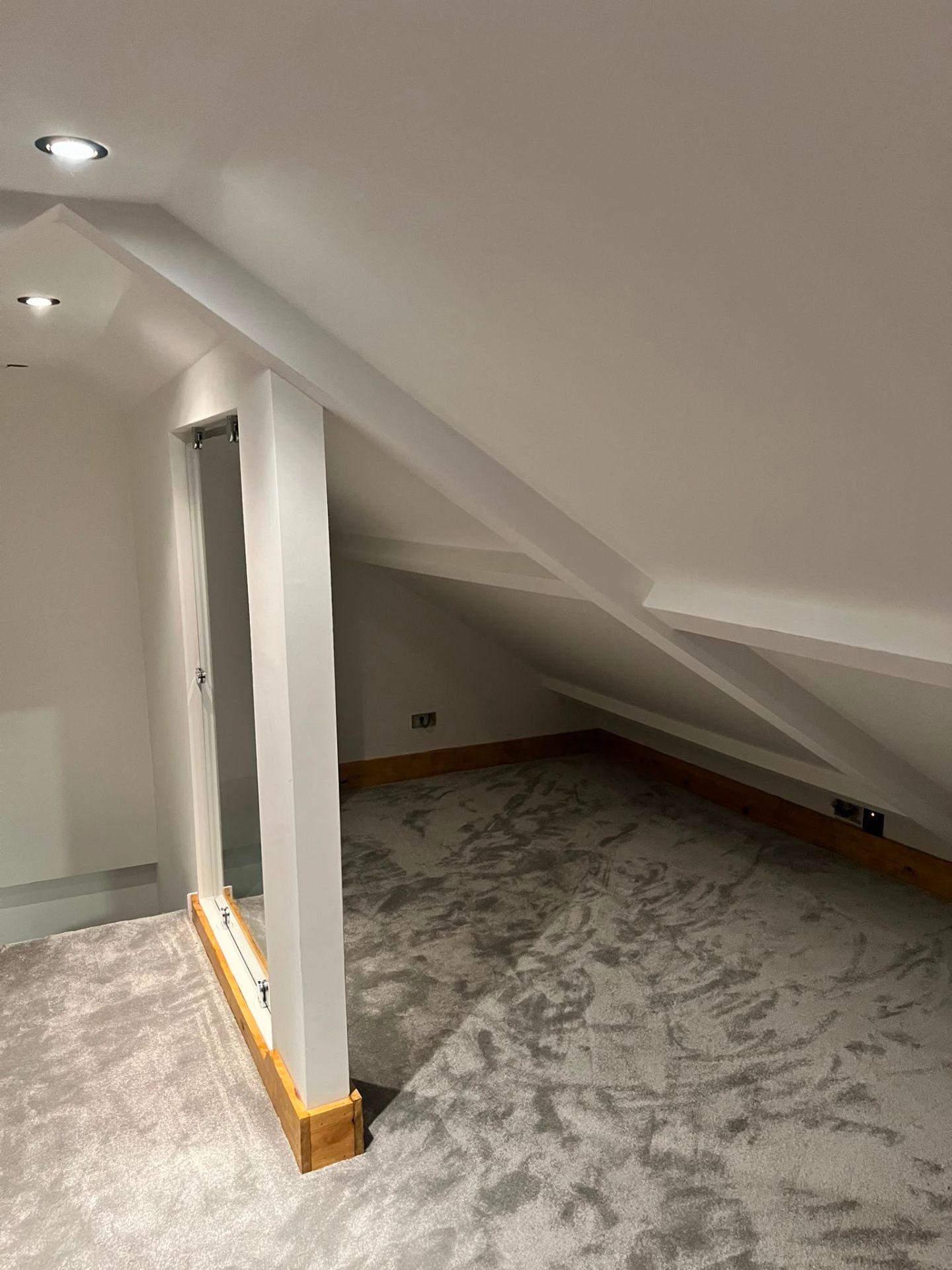 Finished loft conversion space showing sloped ceilings, glass partition, and new grey plush carpeting.