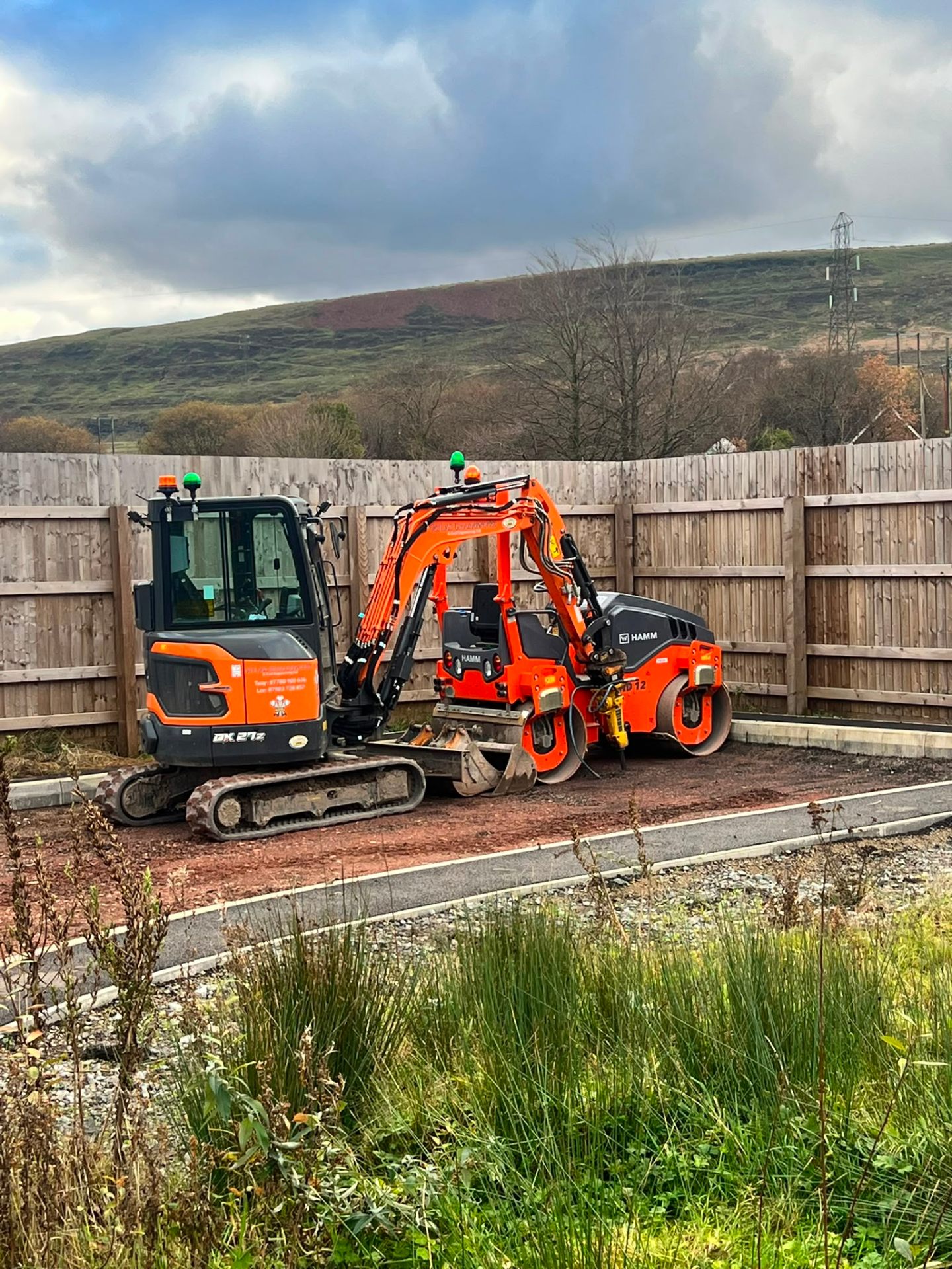 Orange mini digger and roller machinery parked on-site against a wooden fence in South Wales.